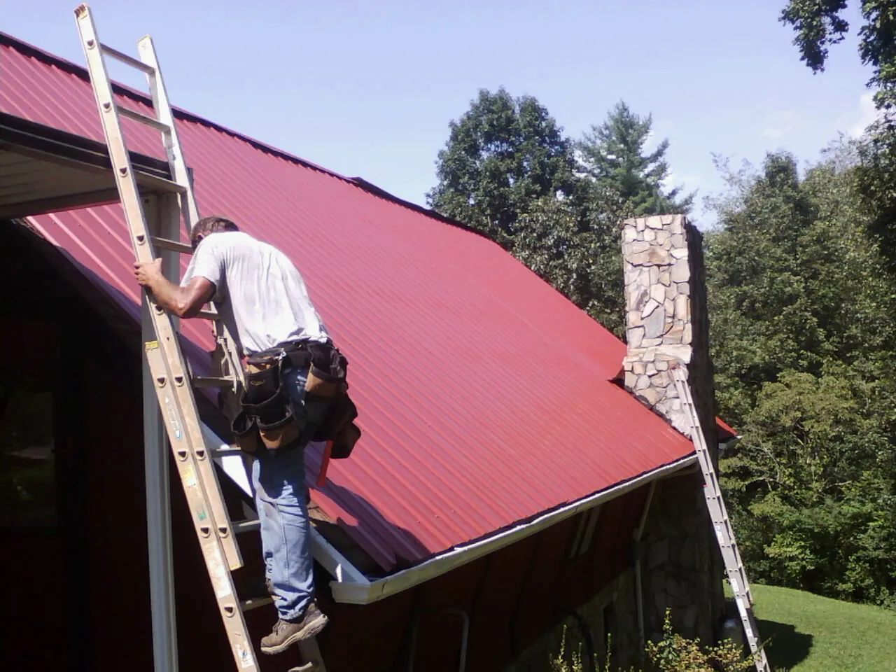 Roofer on a ladder beside a red metal roof and stone chimney — Soffit & Fascia Repair by Roofing Brothers in Hightstown